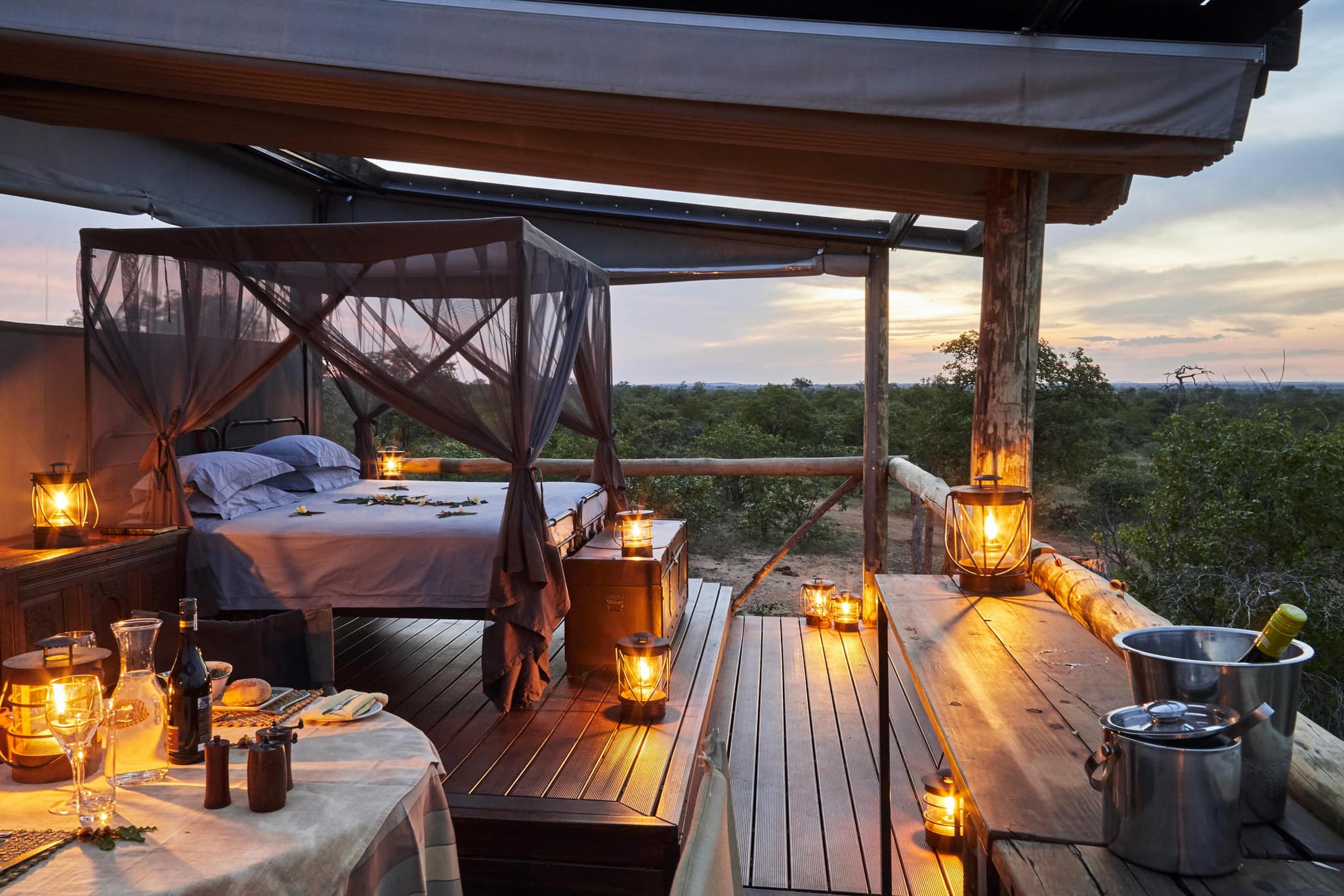 A made-up bed on an open timber deck at dusk, lanterns lit, bush running to the horizon at Motswari