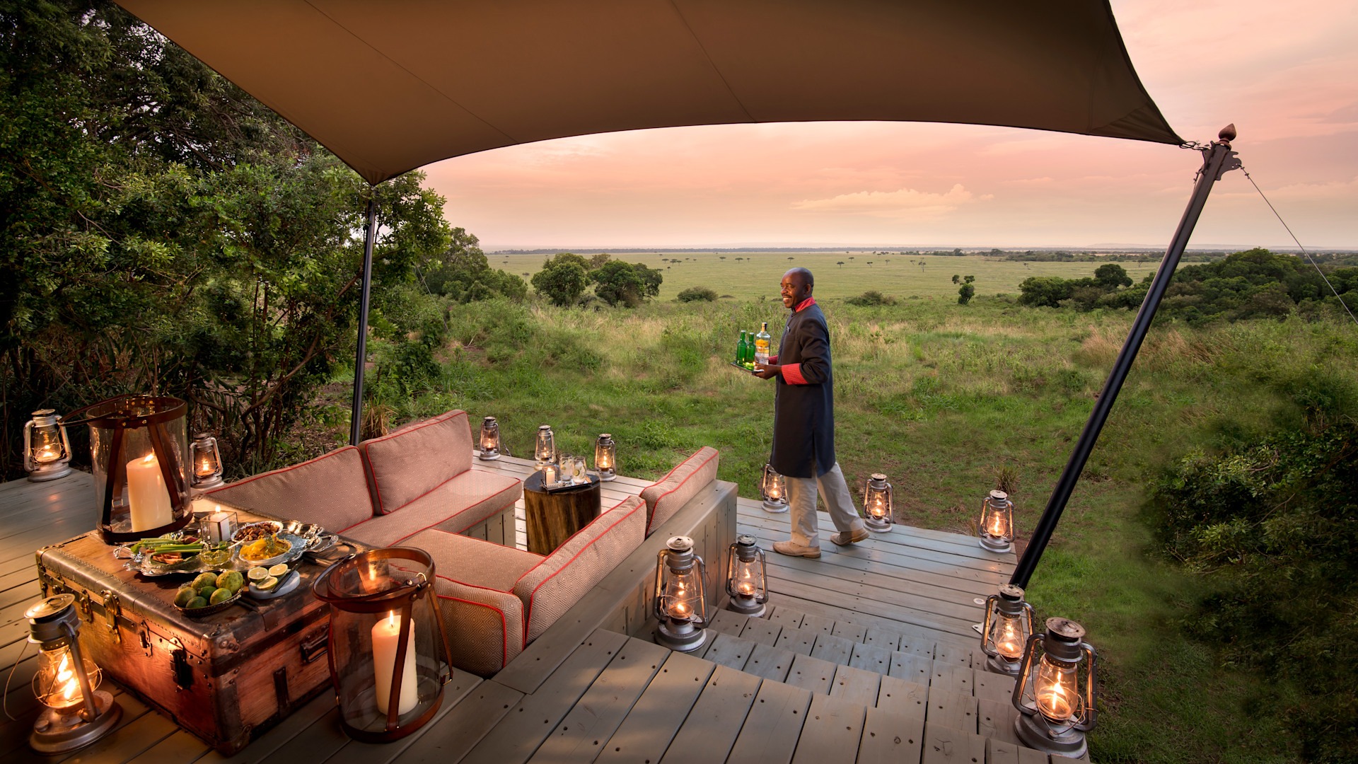 A butler carries drinks across a lantern-lit deck at dusk, the plains of the Masai Mara running to the horizon behind, at &Beyond Bateleur Camp